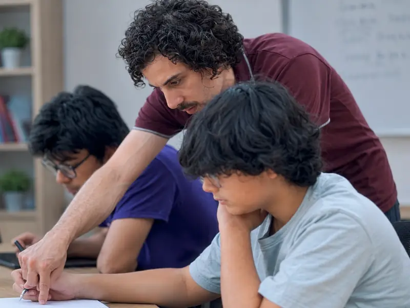 A teacher points to a step in two students’ working at a desk