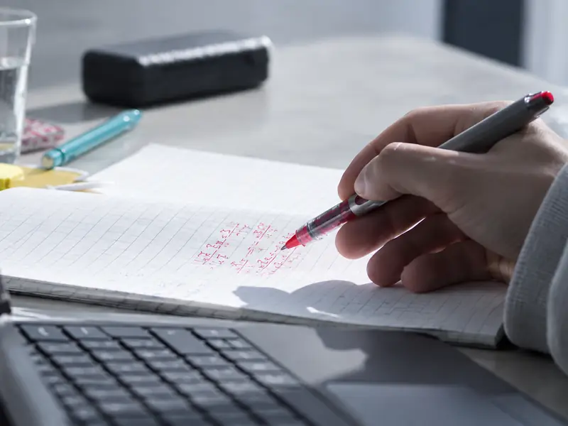 A student writes out working in red pen beside a laptop