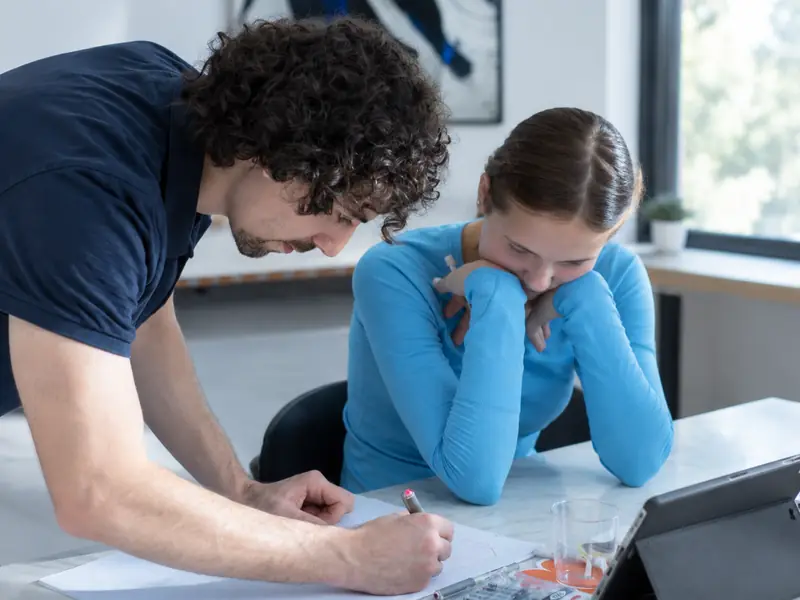 A teacher leans in to check a student’s written working
